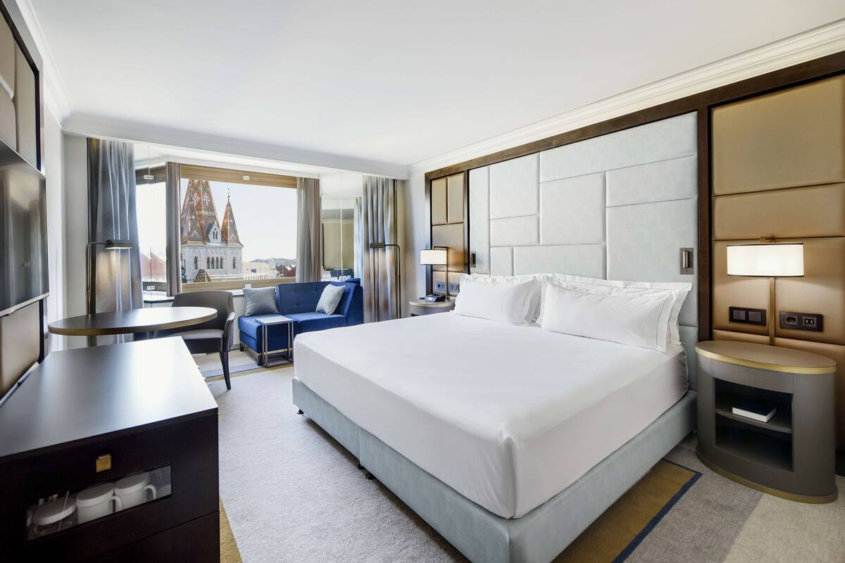 Une chambre d’hôtel avec un grand lit, un bureau, une chaise, une petite table et une vue sur un immeuble surmonté d’une flèche.