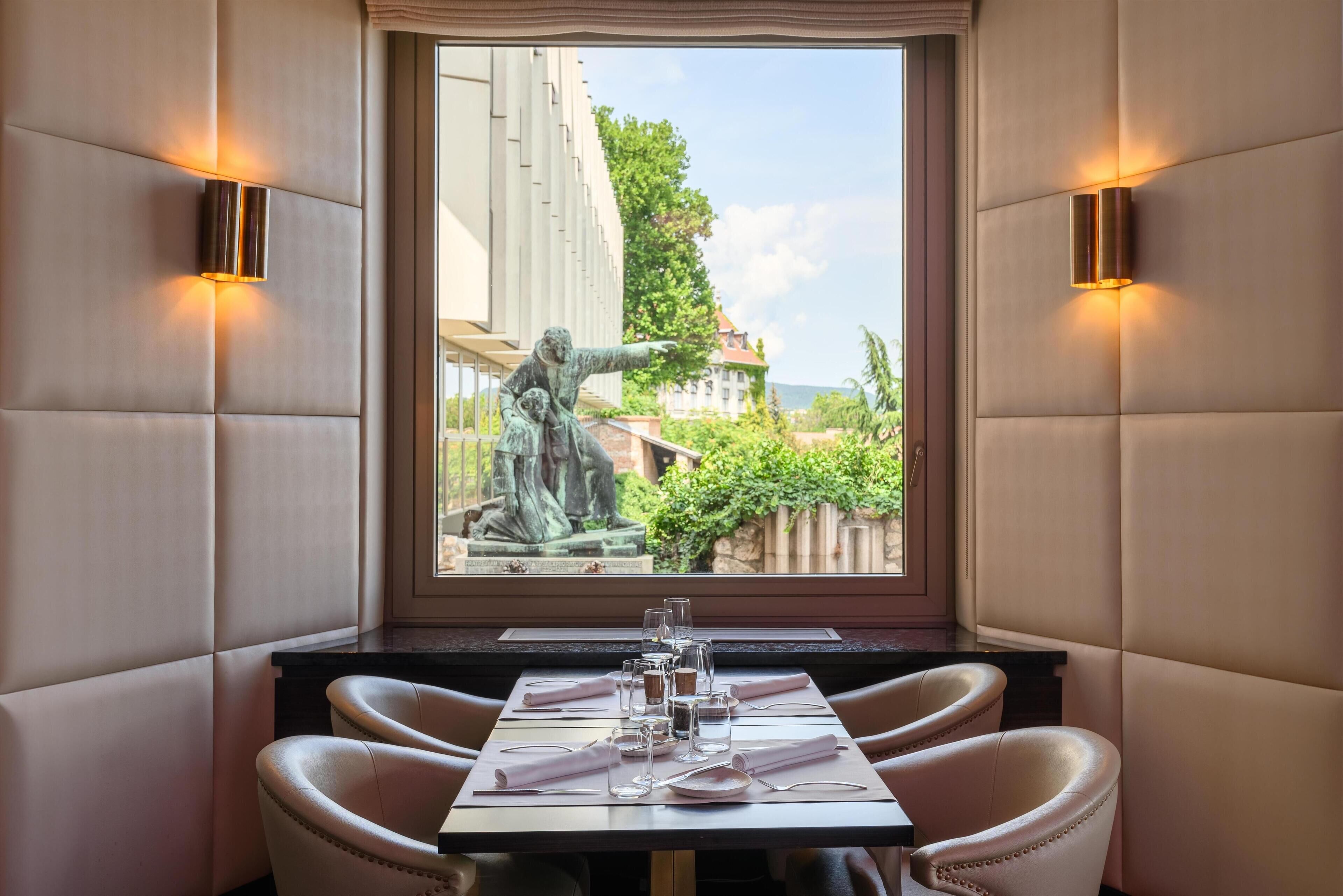 A dining area with a table set for two, a statue in the background, and a view of greenery and buildings outside.
