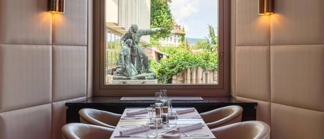 A dining area with a table set for two, a statue in the background, and a view of greenery and buildings outside.
