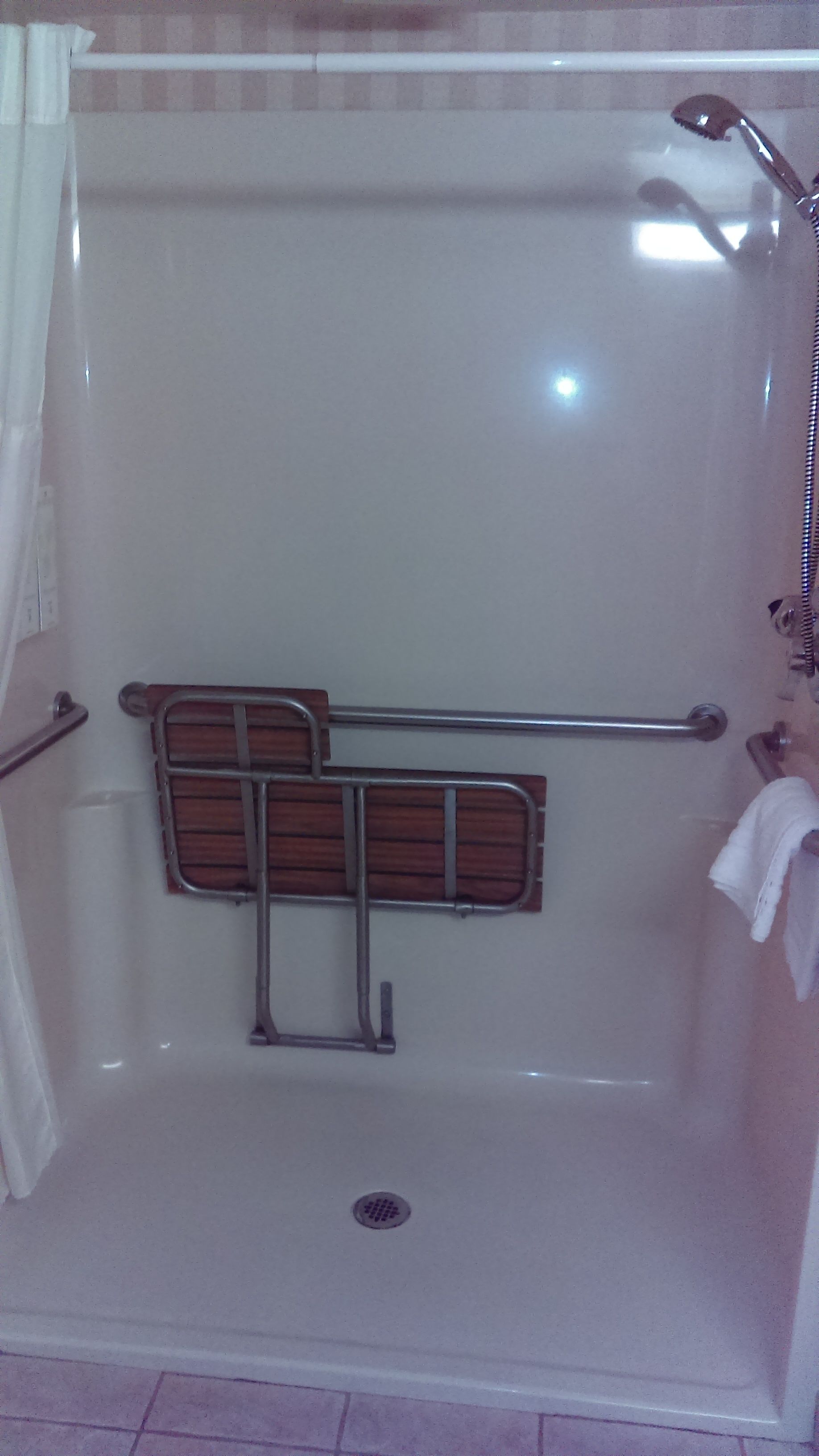 Una doccia con porta in vetro, sedile in legno e barra di sostegno.