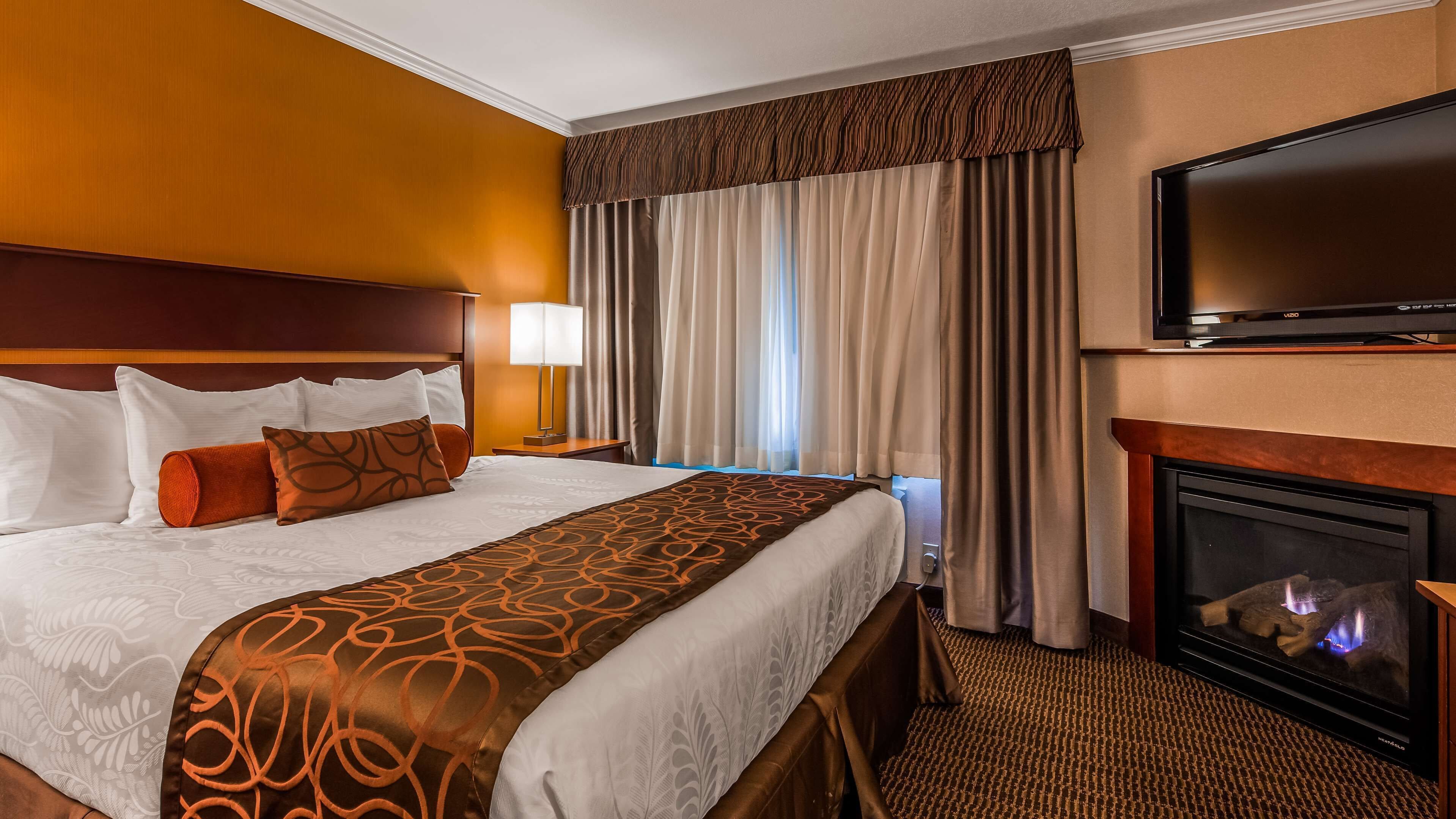 A hotel room with a large bed, a TV, a fireplace, and a window with curtains.