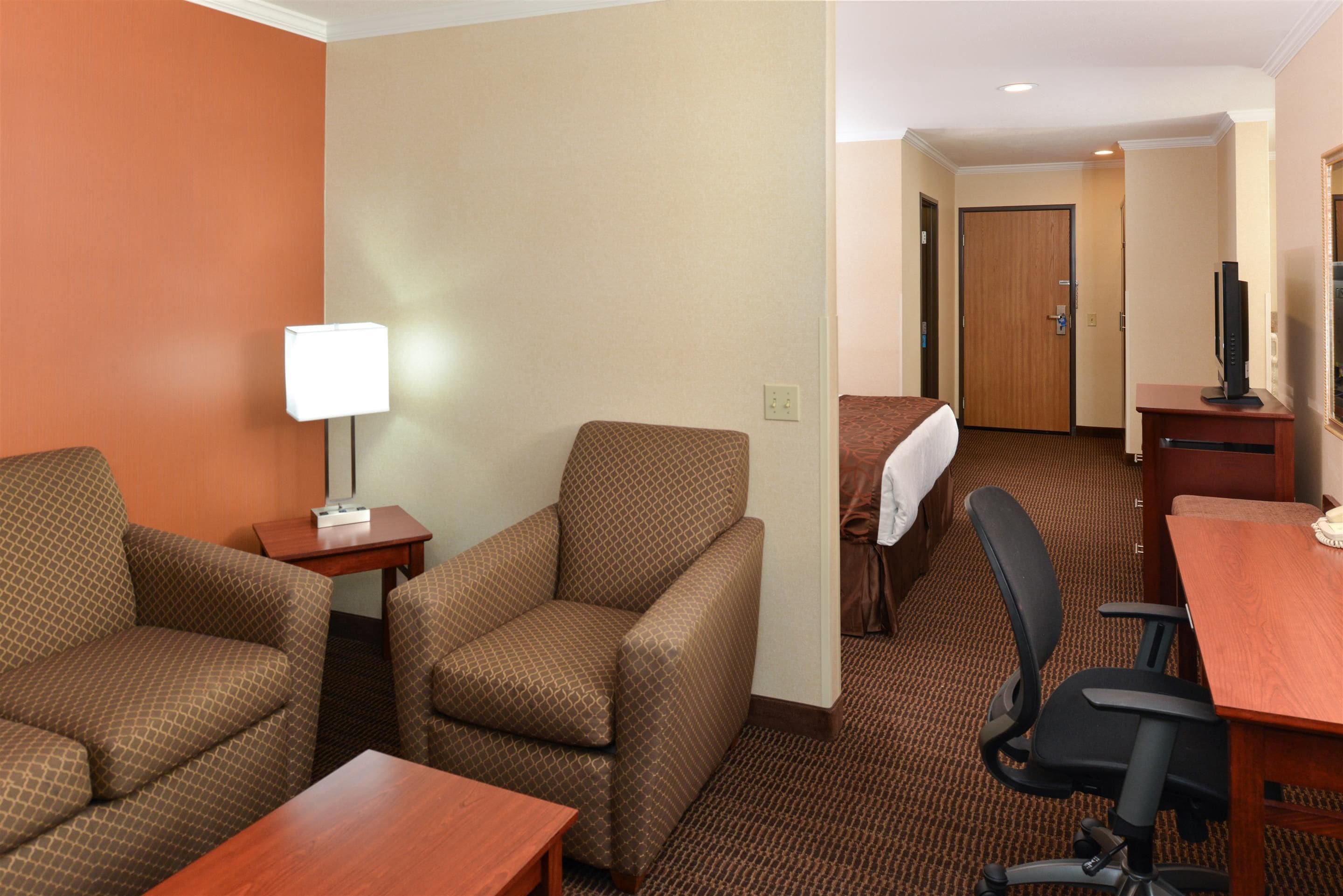 Une chambre d’hôtel avec un canapé marron à motifs, une petite table avec une lampe, un bureau en bois, un fauteuil de bureau noir et un téléviseur posé sur un meuble.