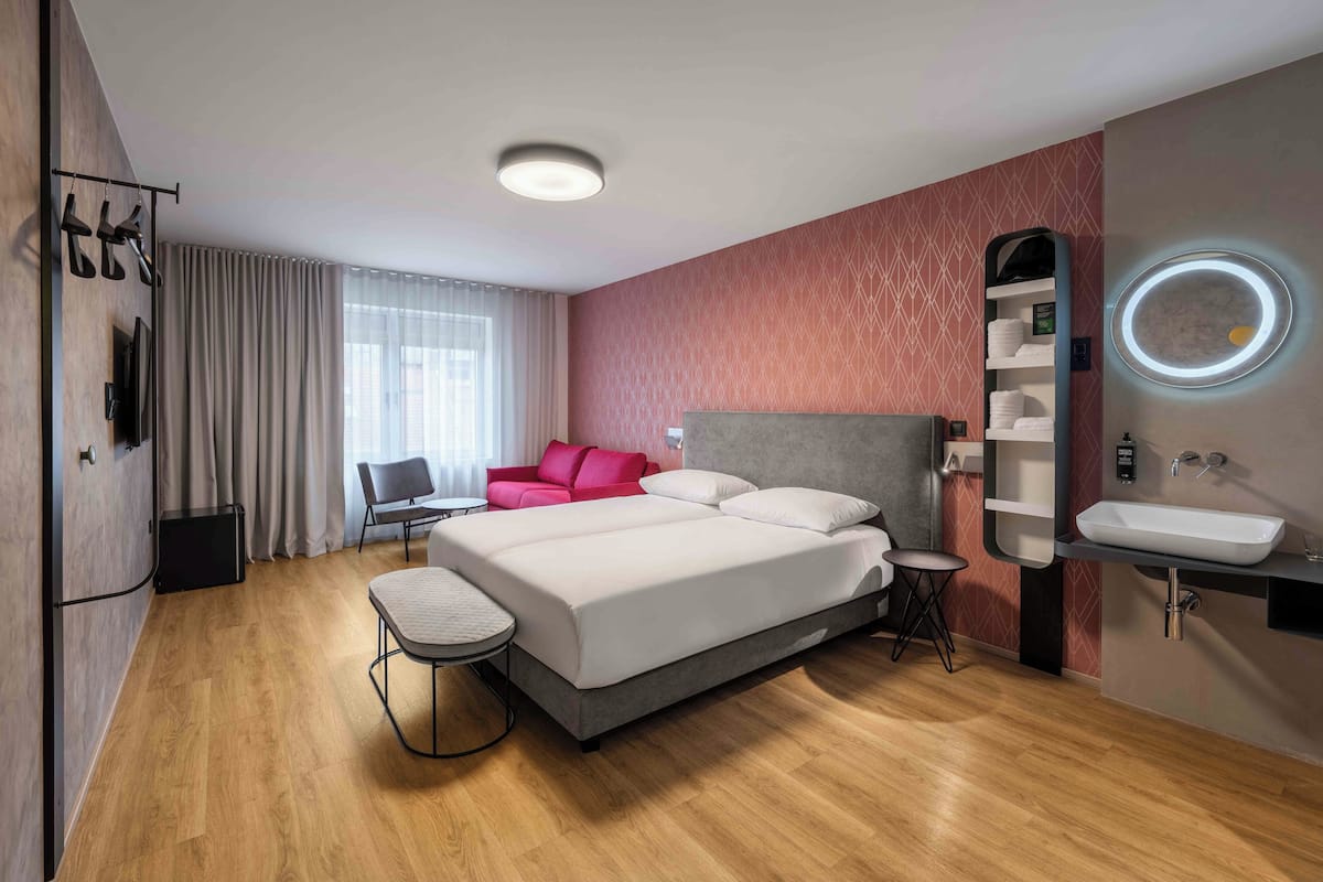 Ein modernes Hotelzimmer mit einem großen Bett, einem Schreibtisch, einem Stuhl, einem kleinen Tisch, einer Couch, einem Spiegel und einem Waschbecken.