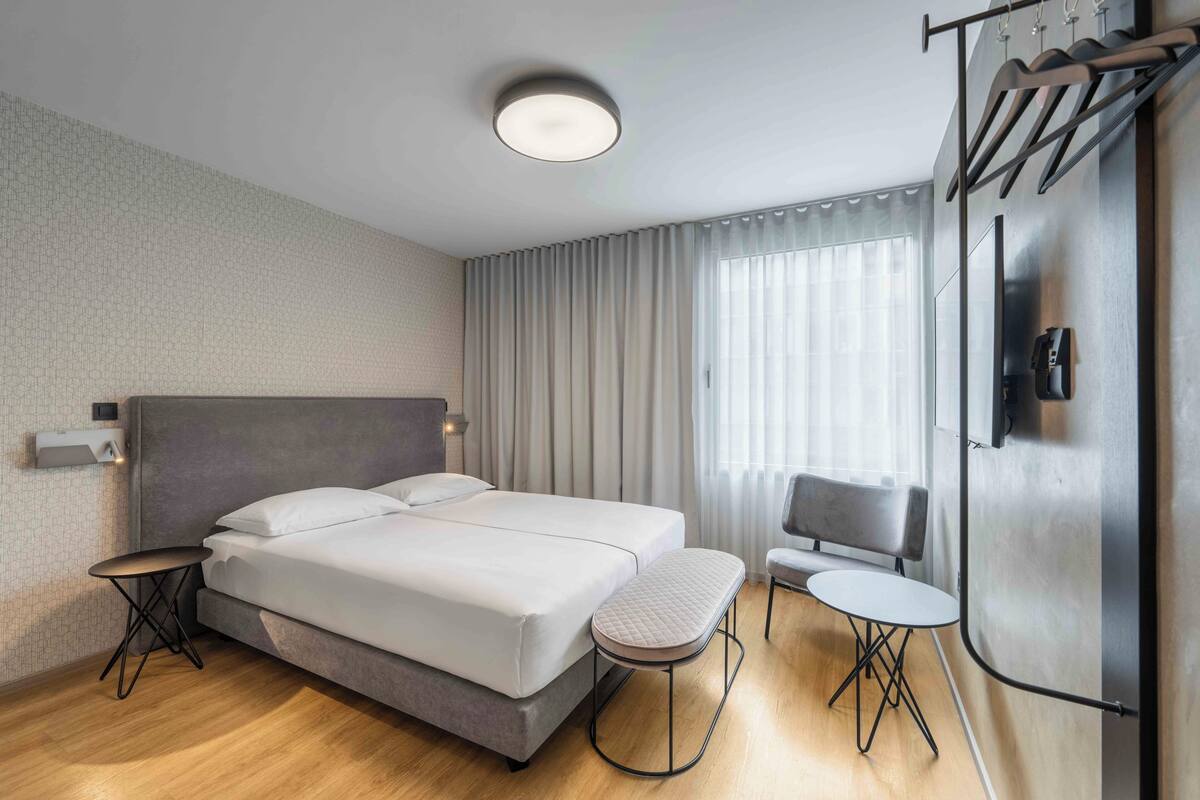 Ein modernes Hotelzimmer mit einem großen Bett, Nachttischen, einer Sitzecke mit Stuhl und kleinem Tisch sowie einem großen Fenster mit Vorhängen.