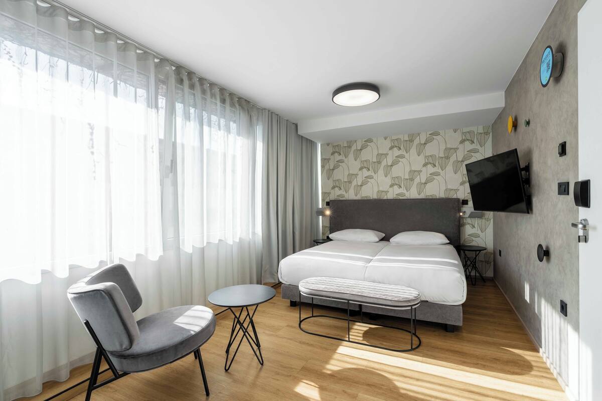 Ein modernes Hotelzimmer mit einem großen Bett, einem Schreibtisch, einem Sessel und einem an der Wand befestigten Fernseher.