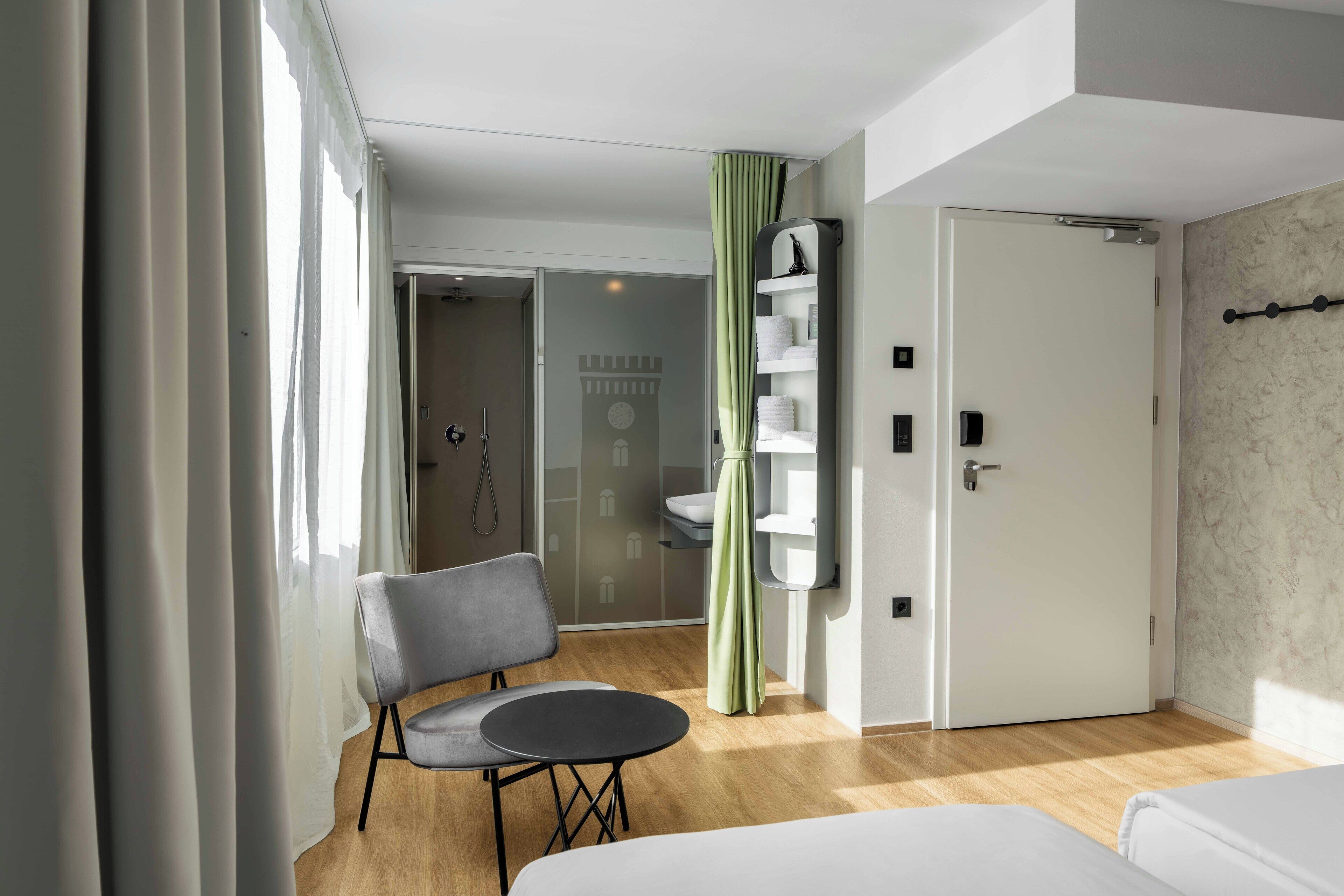 Quarto de hotel com cama, uma cadeira, mesa pequena, banheiro com chuveiro e cortina verde.
