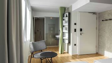 Quarto de hotel com cama, uma cadeira, mesa pequena, banheiro com chuveiro e cortina verde.