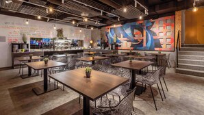 Een modern restaurantinterieur met meerdere eettafels en stoelen, een keukenhoek, en een kleurrijke abstracte muurschildering.