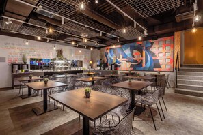 A modern restaurant interior with multiple dining tables and chairs, a kitchen area, and a colorful abstract mural on the wall.