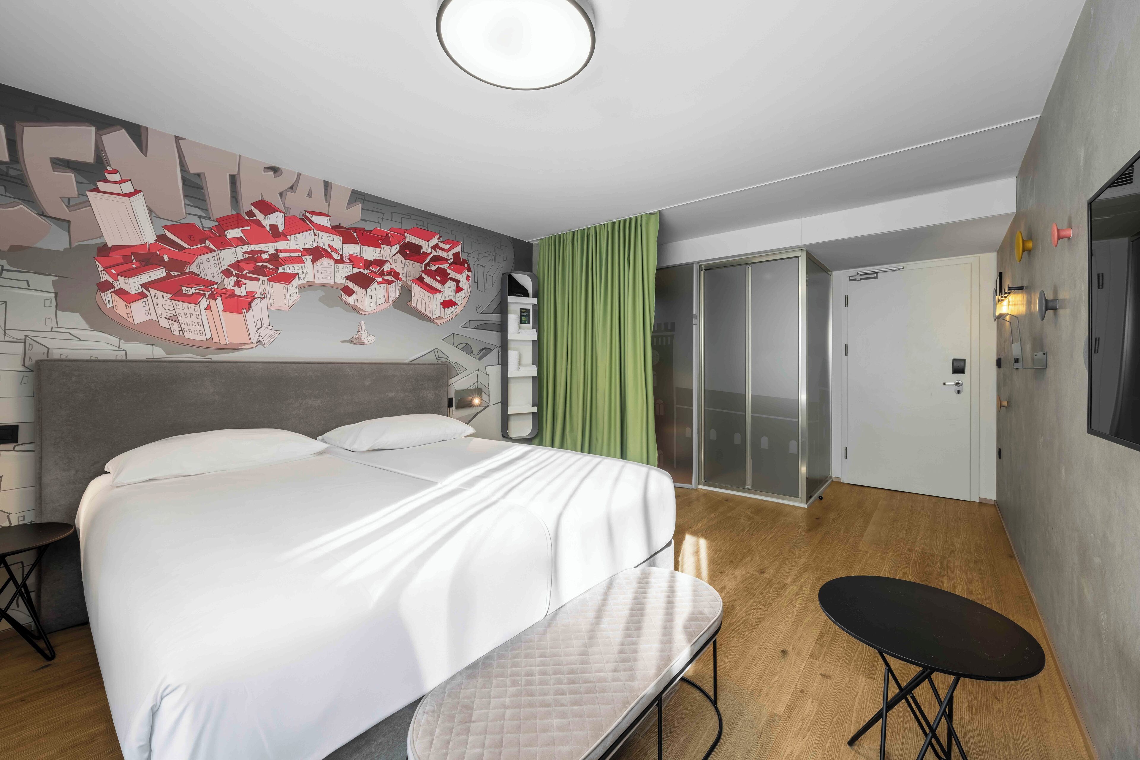 A modern hotel room with a large bed, a small table, a green curtain, and a cityscape mural on the wall.