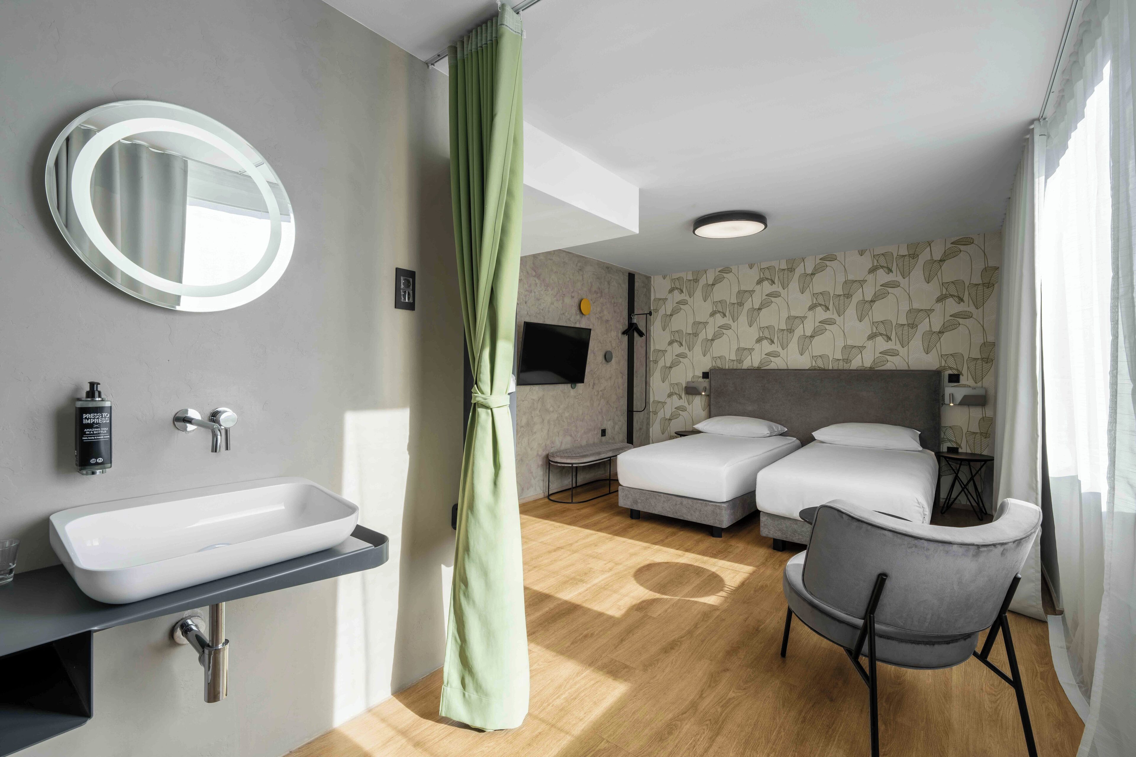 A modern hotel room with a large bed, a bedside table, a chair, a round mirror, a sink, and a shower curtain.