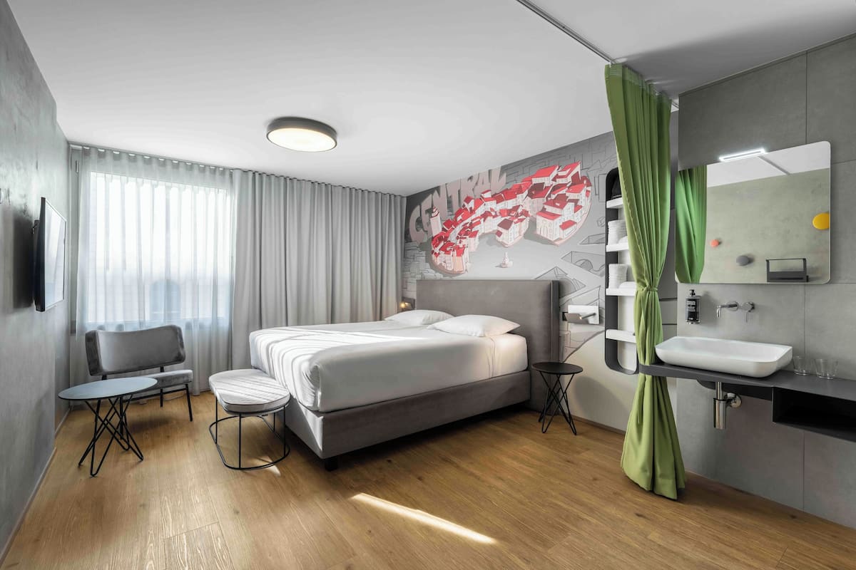 Ein modernes Hotelzimmer mit einem großen Bett, einem kleinen Tisch, einem Sessel, einer Couch, einem Badezimmer mit Waschbecken und Spiegel sowie einem Wandbild.