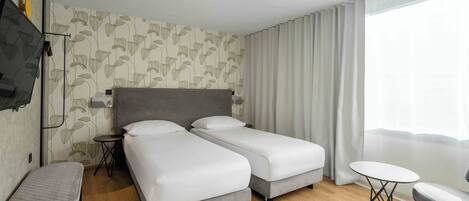 Habitación de hotel con dos camas individuales, una mesita, una silla, televisor y ventana con cortinas.