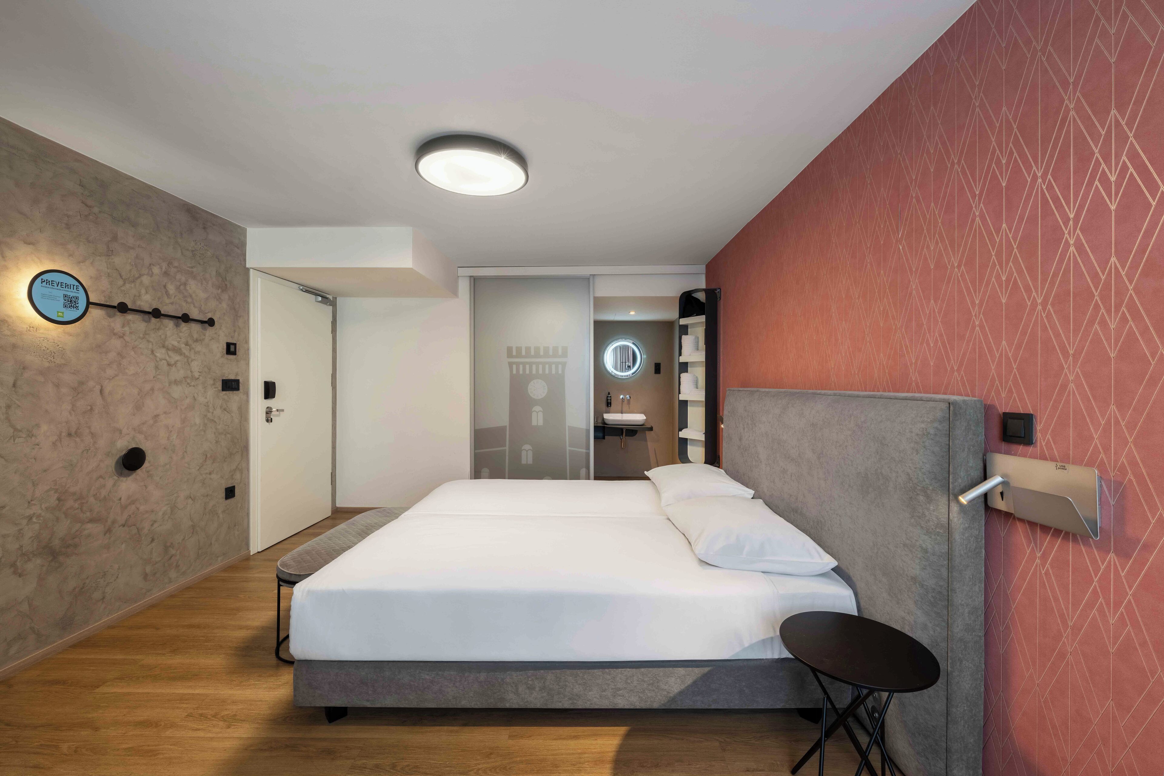 Une chambre d’hôtel moderne avec un grand lit, une table de chevet, une étagère intégrée et une salle de bain visible à travers une porte ouverte.