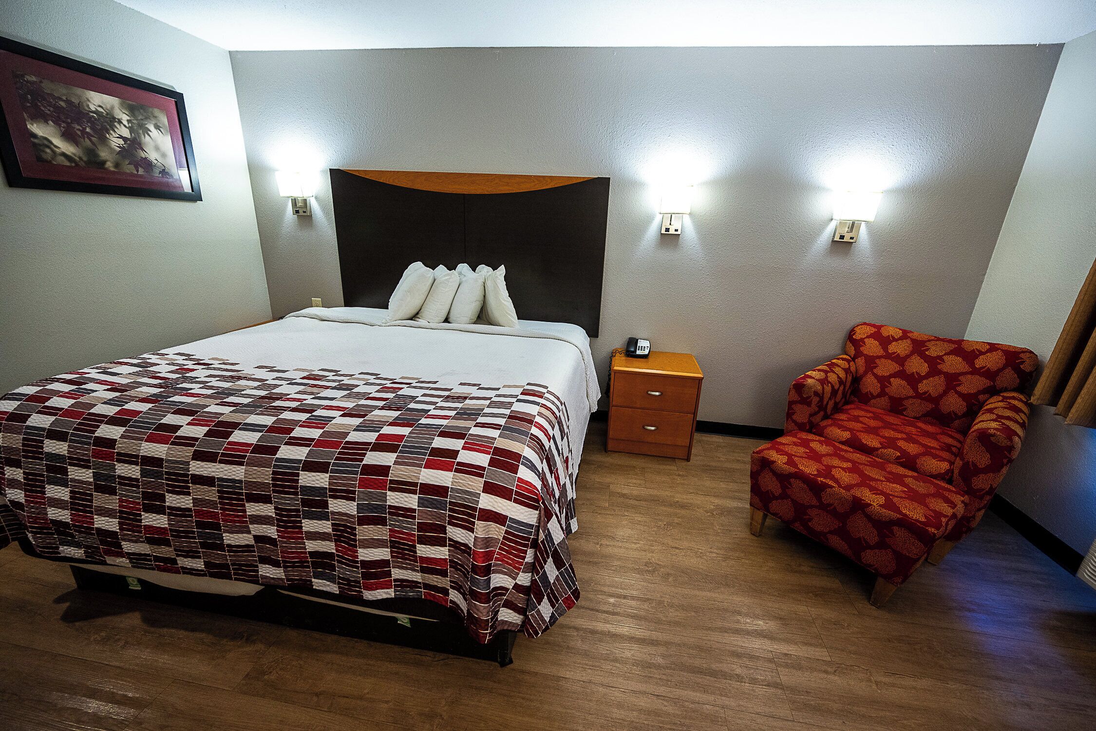 A hotel room with a bed, a nightstand, a chair, and a wall-mounted light fixture.