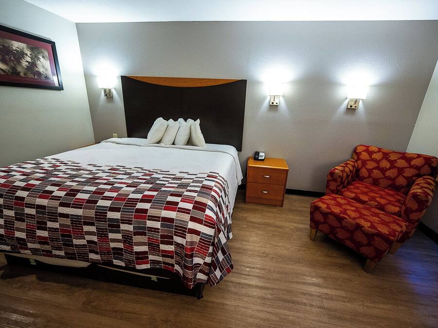 A hotel room with a bed, a nightstand, a chair, and a wall-mounted light fixture.