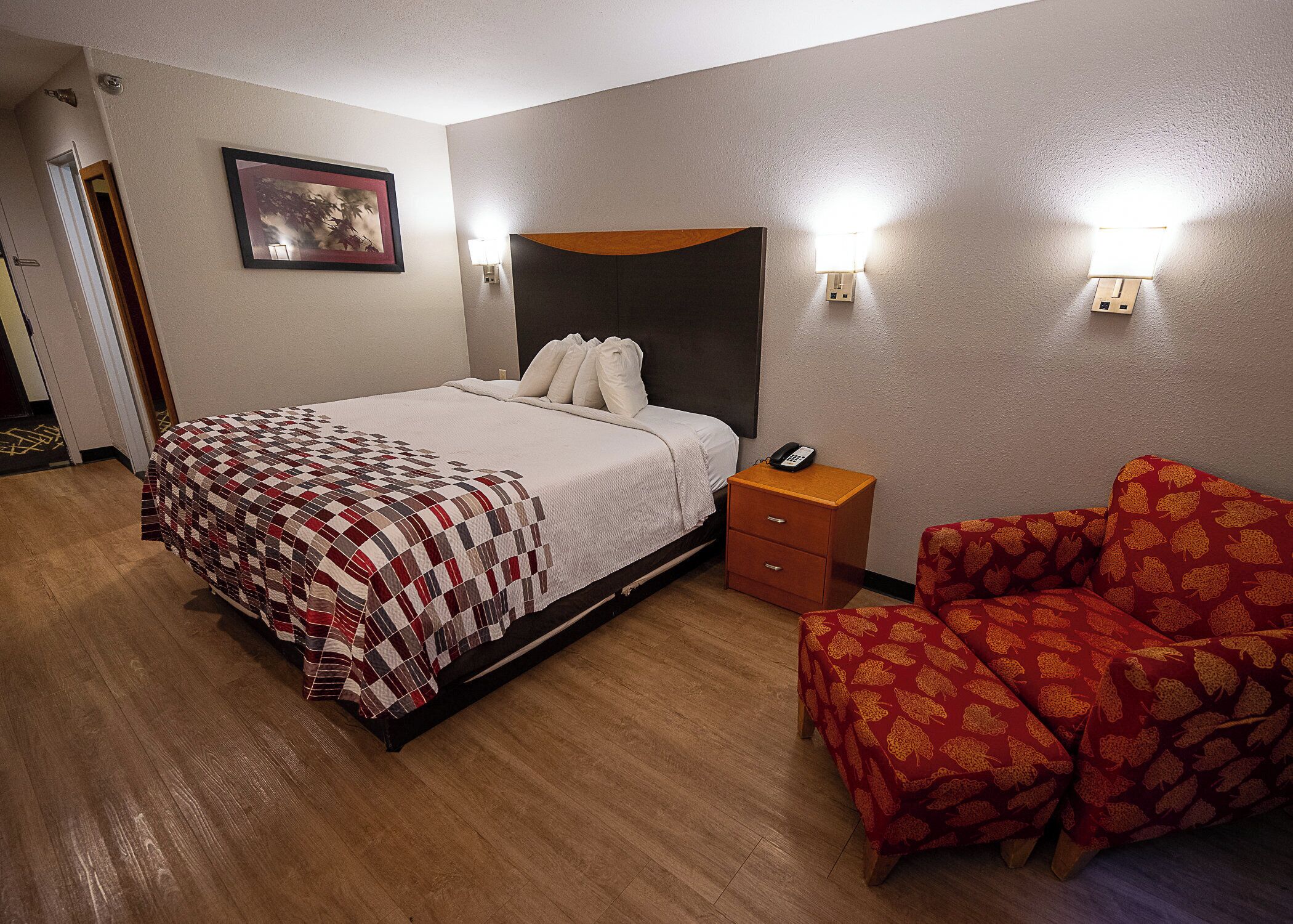 A hotel room with a bed, bedside table, chair, and wall-mounted lights.