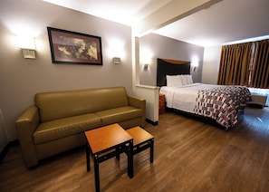 A hotel room with a sofa, a small table, a bed with a patterned blanket, and a painting on the wall. - Red Roof Inn & Suites Danville, IL (Danville)