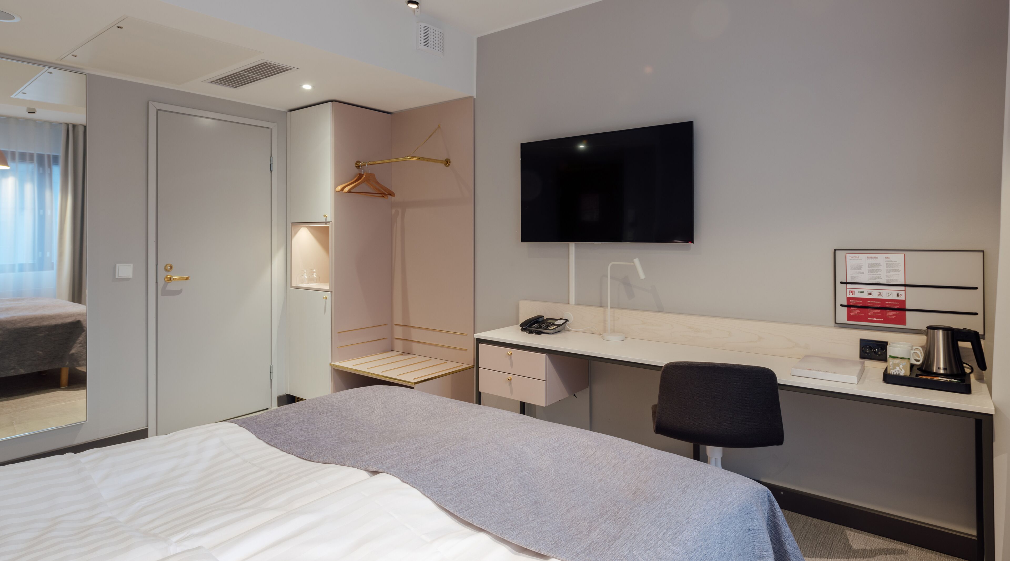 Hotellihuone, jossa on sänky, työpöytä, tuoli, televisio, naulakko, peili ja pieni hylly, jonka päällä on puhelin ja vedenkeitin.