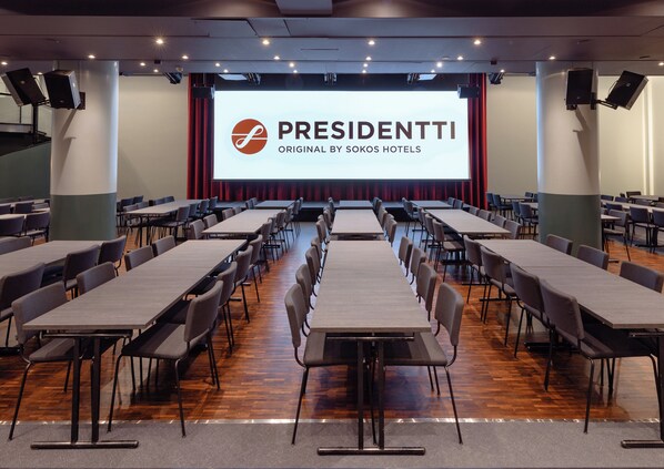 Un salón de conferencias con filas de mesas y sillas, una gran pantalla mostrando el logotipo "PRESIDENTTI ORIGINAL BY SOKOS HOTELS", y un escenario con cortinas.