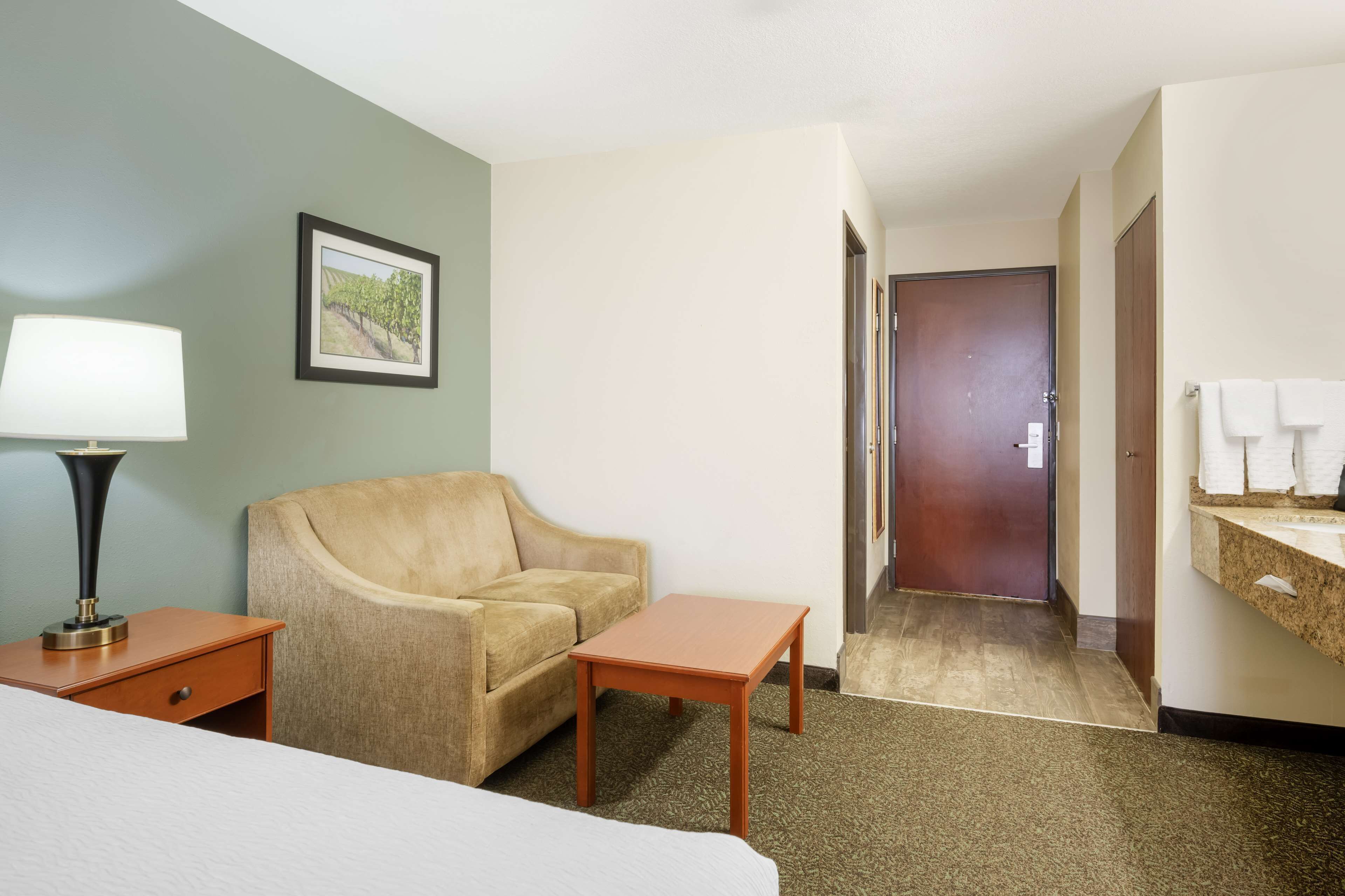 Habitación de hotel con cama, sofá, una mesita, lámpara, un cuadro en la pared y baño con lavamanos y toallero.