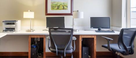Un espacio de oficina moderno con dos escritorios, cada uno con una computadora y una silla. Hay dos lámparas fijadas a la pared, un cuadro de un paisaje enmarcado e impresoras a la izquierda.