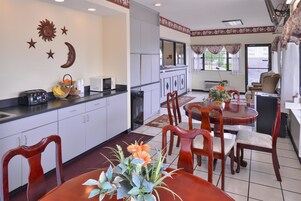 A hotel room with a kitchen area, dining table, and chairs. There are decorative wall elements and a window with curtains.