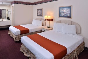 Quarto de hotel com duas camas, uma mesa de cabeceira com abajur, um quadro na parede e um banheiro visível ao fundo.