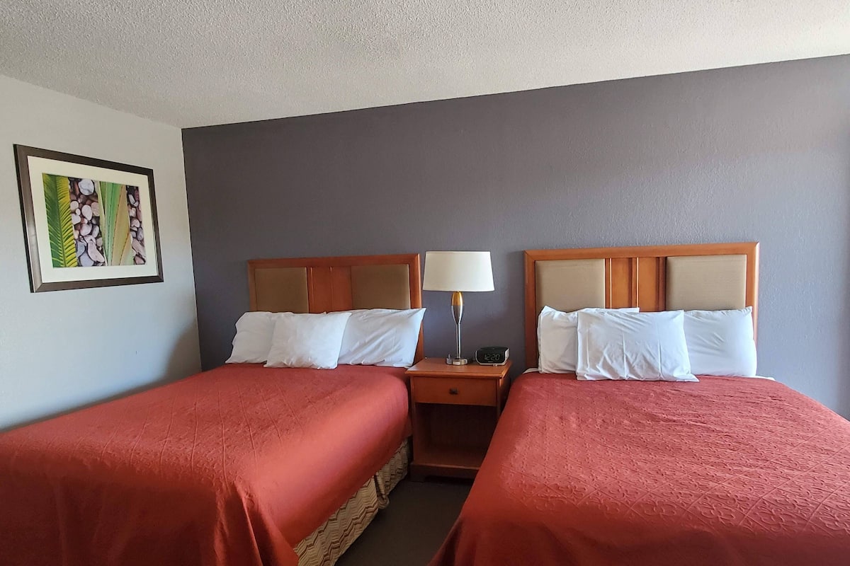 Et hotelværelse med to senge, hver med en trægavl og hvide sengetøj. Et natbord med lampe og ur står placeret mellem sengene. Et indrammet billede hænger på væggen.