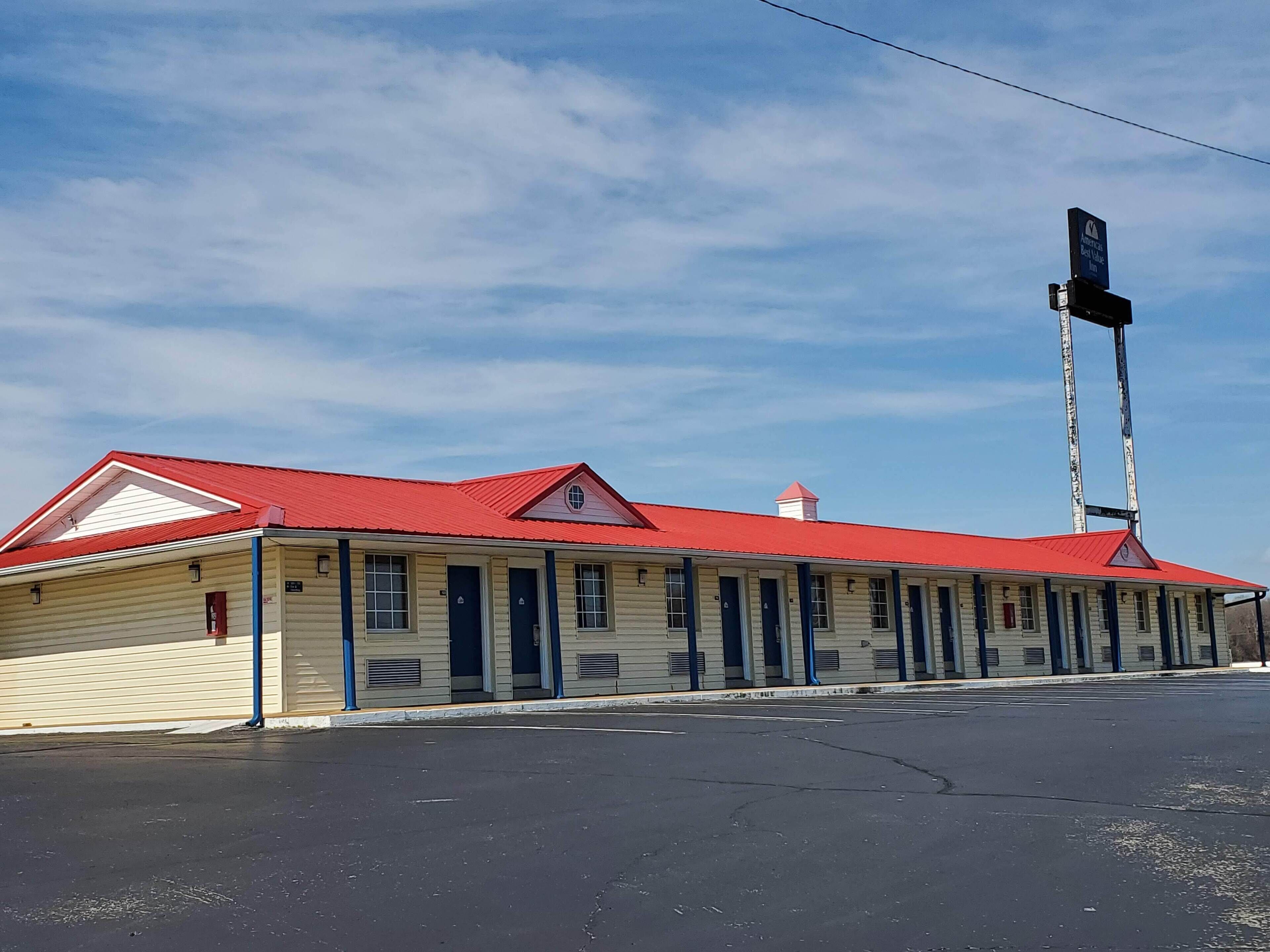 A two-story motel with a red roof and beige siding, featuring a row of single rooms with dark blue doors.