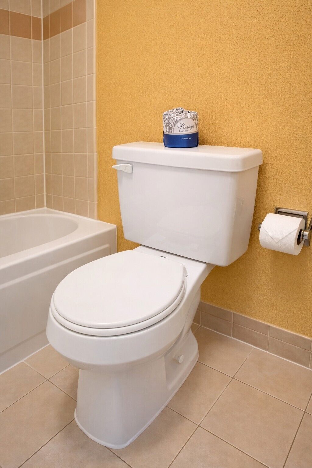 A white toilet with a tank and lid, a toilet paper holder on the wall, and a bathtub in the background.