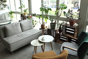 Una sala moderna con un sofá, una mesa de centro y un sillón. Hay varias plantas en macetas y vistas al exterior a través de amplios ventanales.