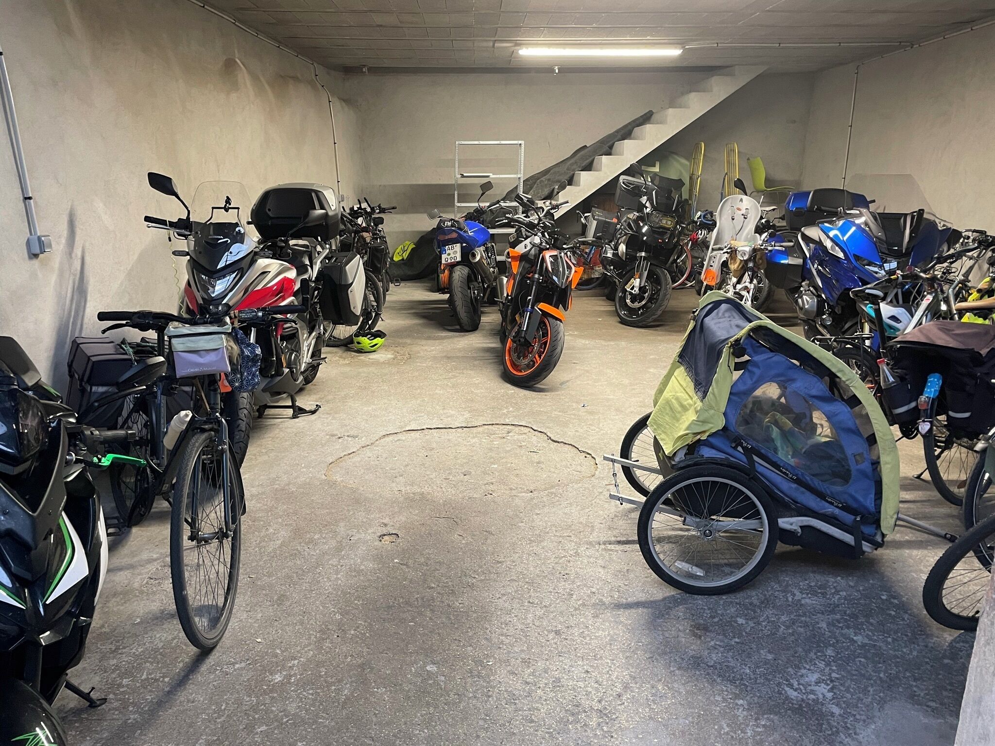 Un garaje para estacionamiento con varias motocicletas y bicicletas, piso de concreto y una escalera al fondo.