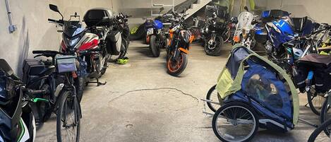 A parking garage with multiple motorcycles and bicycles, a concrete floor, and a staircase in the background.