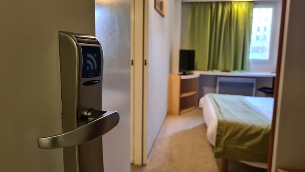 Habitación de hotel con puerta equipada con cerradura digital, cama, escritorio con computadora y ventana con cortinas.