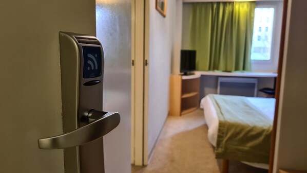 Quarto de hotel com porta equipada com fechadura digital, uma cama, uma escrivaninha com computador e uma janela com cortinas.