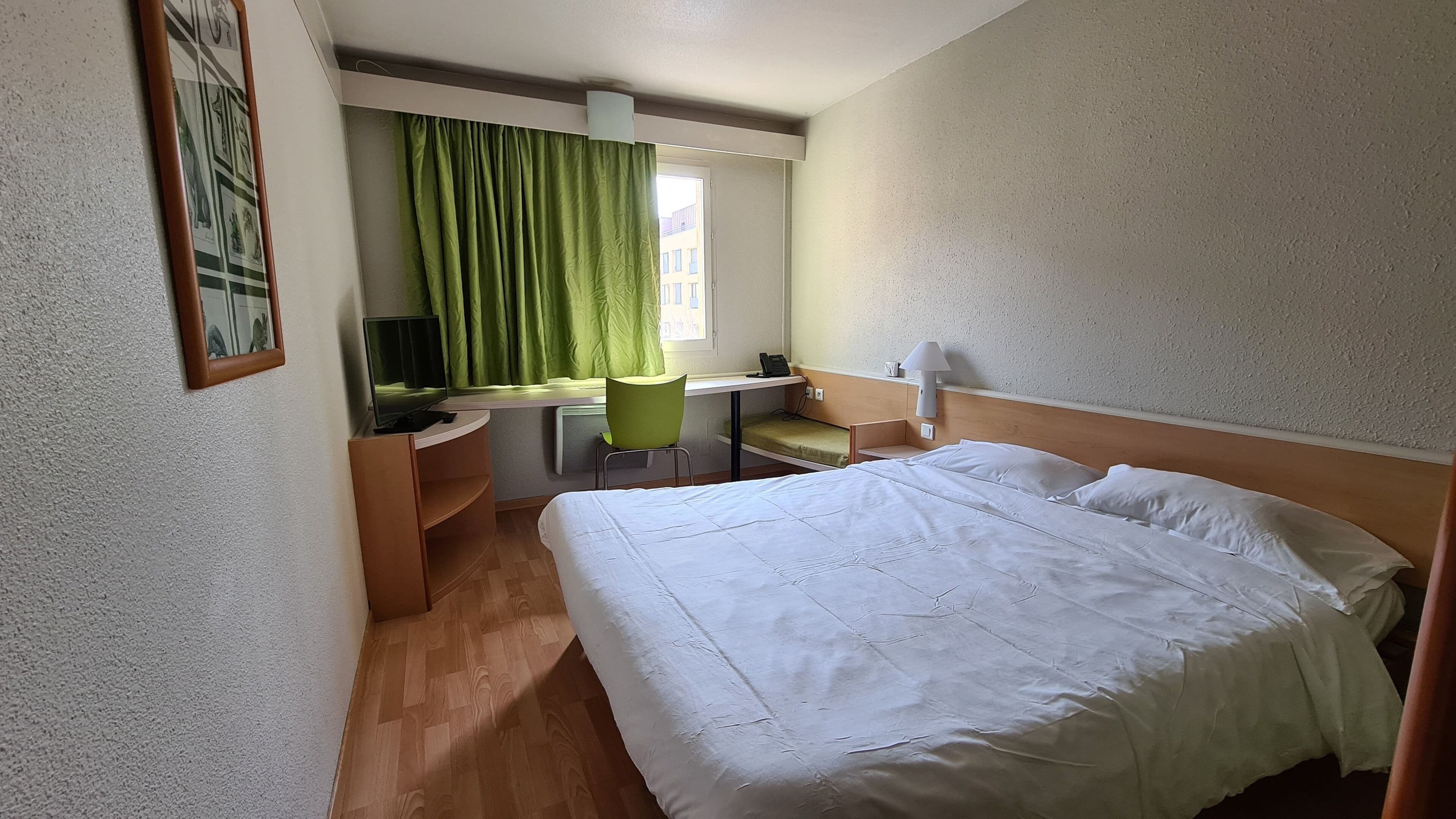 Une chambre d’hôtel avec un lit, un bureau et une chaise, une télévision et une fenêtre avec des rideaux verts.