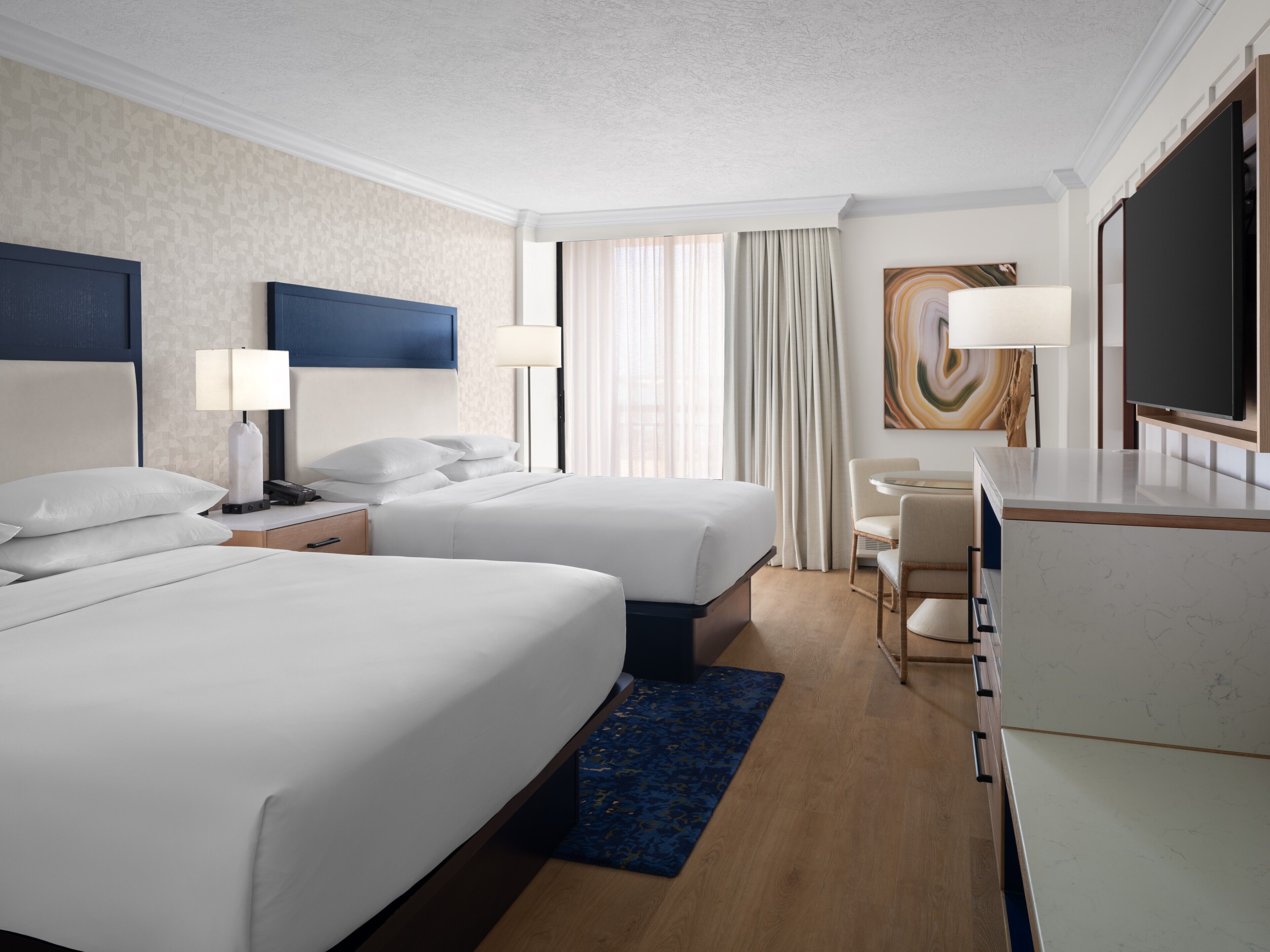 A hotel room with two beds, a desk, a chair, a TV, and a large window with curtains.