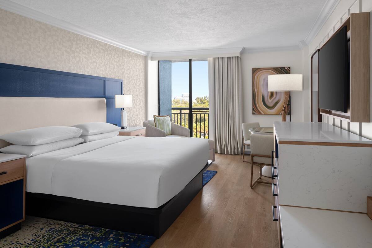 A hotel room with a large bed, a desk, a TV, and a balcony with a view.