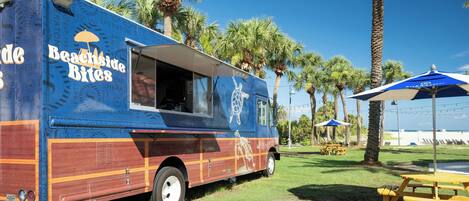 A beachside food truck with a turtle design, parked in a grassy area with palm trees and picnic tables.