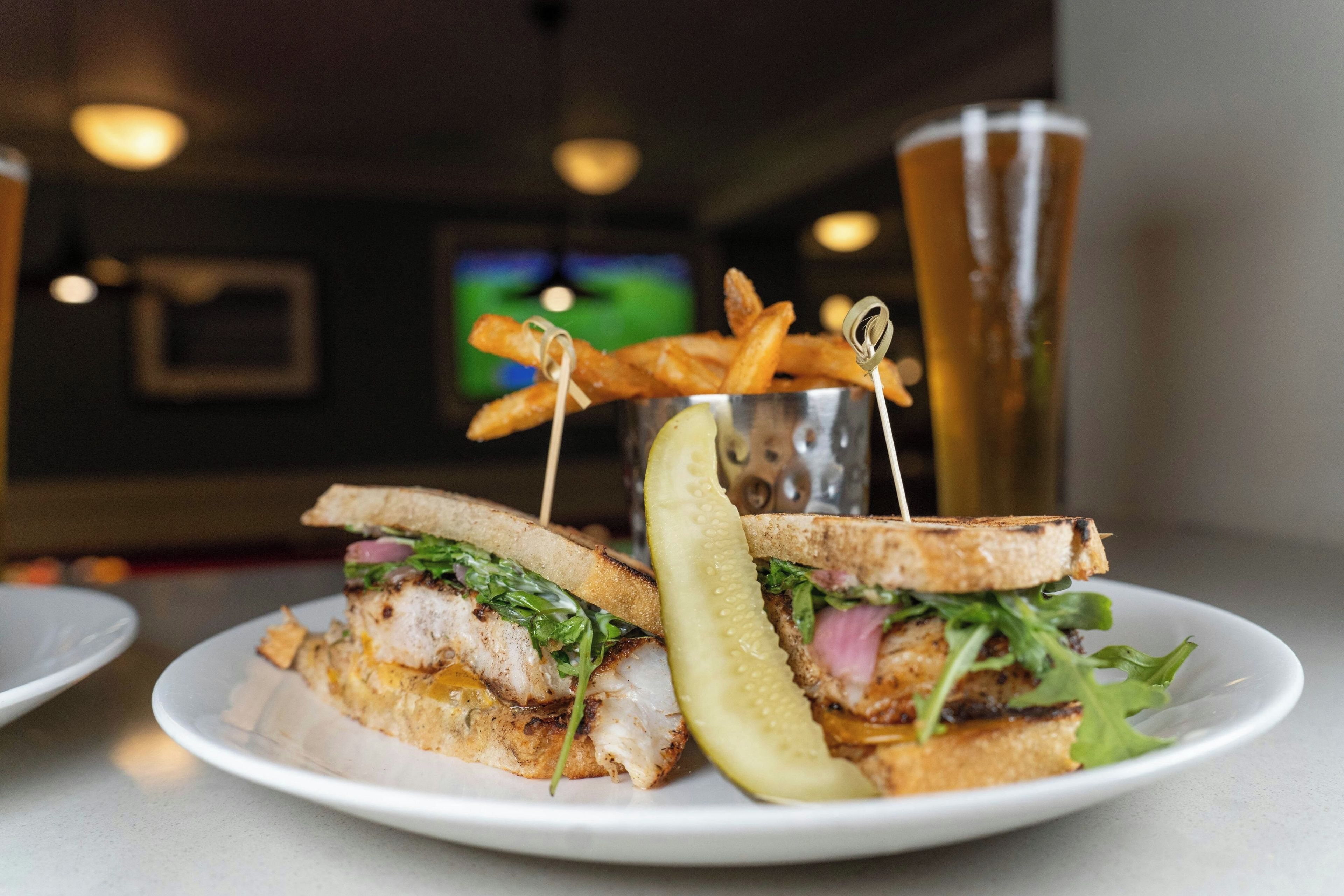 A sandwich with a pickle on a plate, a glass of beer, and a basket of fries in the background.