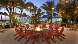 A fire pit surrounded by red Adirondack chairs in an outdoor patio area with palm trees and umbrellas. - Clearwater Beach Sheraton Resort on Sand Key (Clearwater Beach)