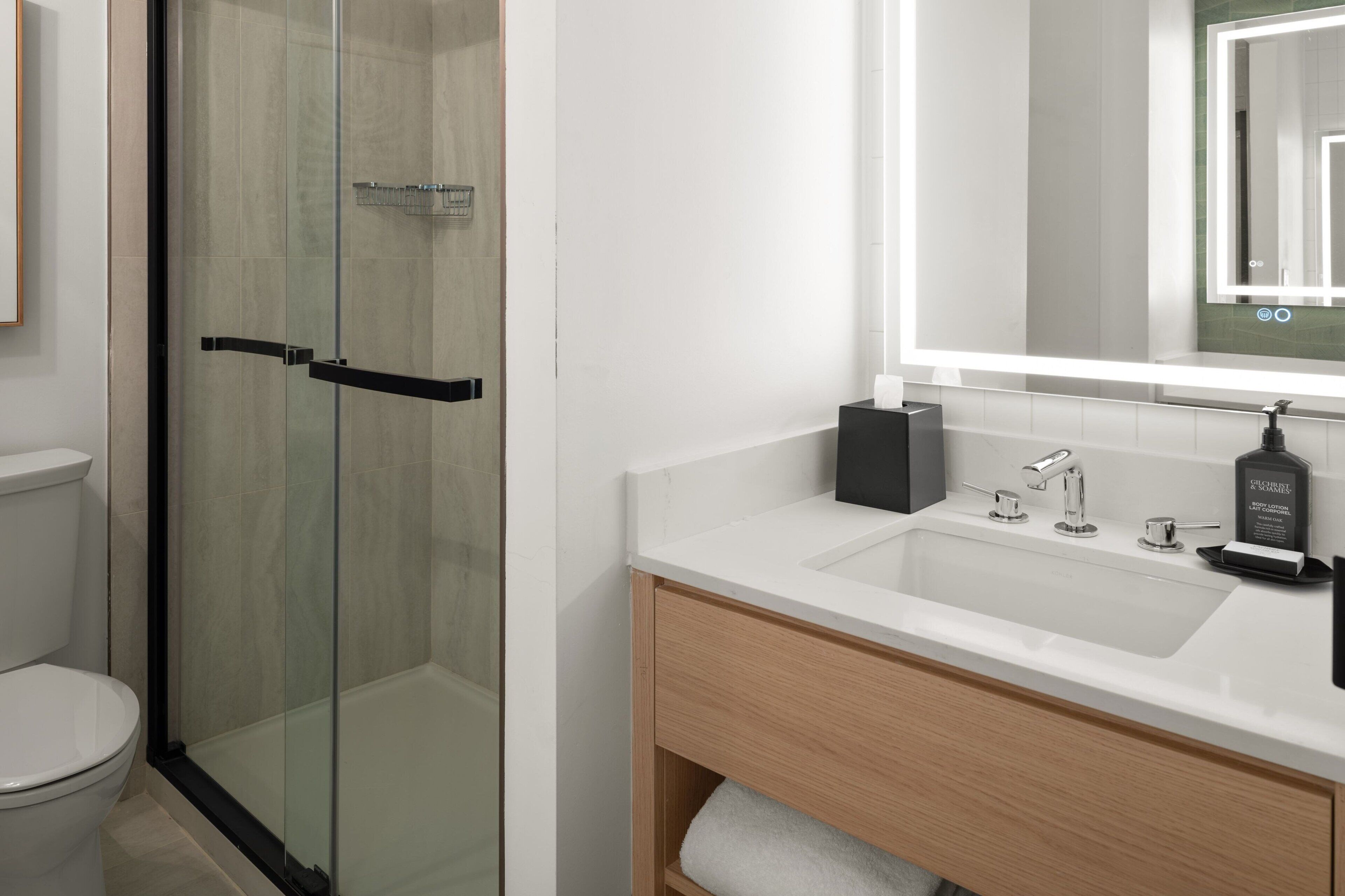 Un baño moderno con ducha envuelta en vidrio, un lavamanos blanco con espejo arriba y un mueble de tocador de madera.