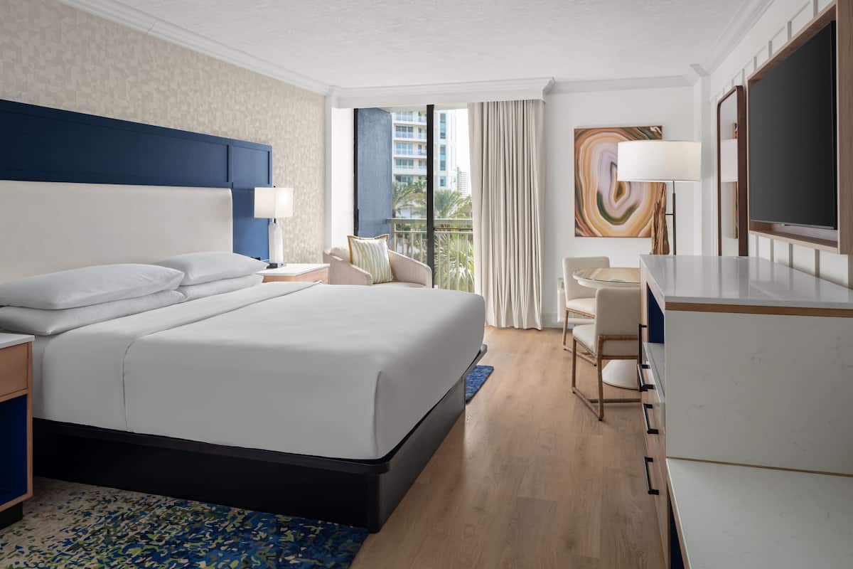 A modern hotel room with a large bed, a desk, a chair, a TV, and a balcony with a view of the city.