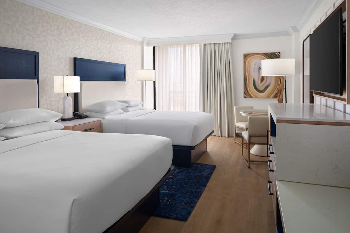 A hotel room with two beds, a desk, a chair, a TV, and a large window with curtains.