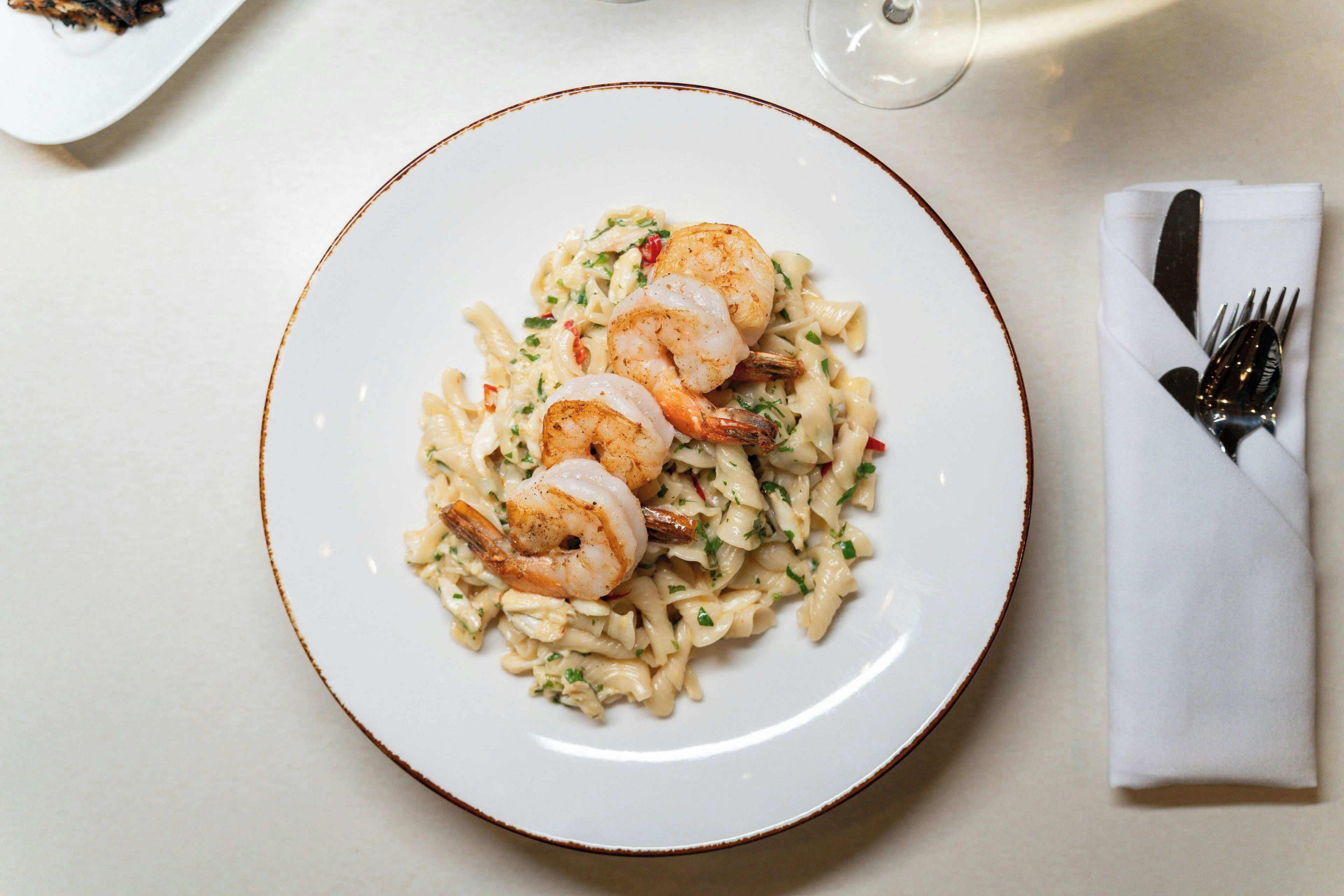 A plate of pasta with shrimp on a white plate with a brown rim, accompanied by a fork and a napkin on a table.