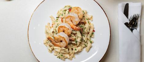 A plate of pasta with shrimp on a white plate with a brown rim, accompanied by a fork and a napkin on a table.