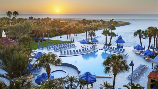 A resort pool area with sun loungers and umbrellas, surrounded by palm trees and a view of the ocean at sunset. - Clearwater Beach Sheraton Resort on Sand Key (Clearwater Beach)