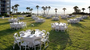 An outdoor dining area with round tables and white chairs, set on a grassy lawn with palm trees and a building in the background. - Clearwater Beach Sheraton Resort on Sand Key (Clearwater Beach)