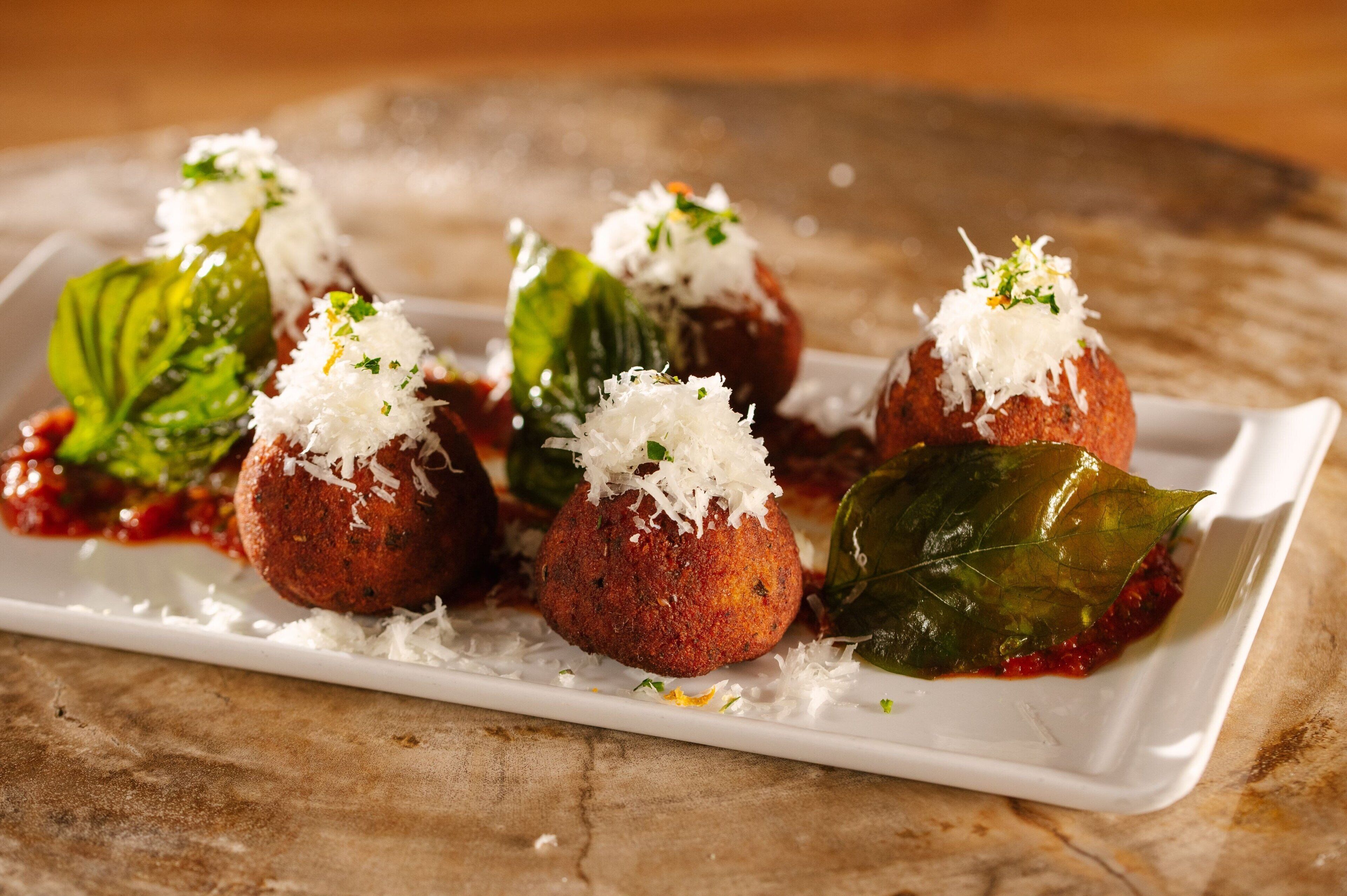 A white rectangular plate with several round, golden-brown fried items topped with shredded cheese and green leaves, served on a textured surface.
